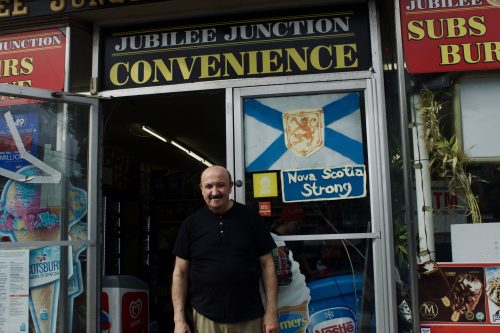 A man wearing black, smiling outside his store, the sign behind him says Jubilee Junction Convenience and there is a photo of Nova Scotia's flag with a title Nova Scotia Strong.