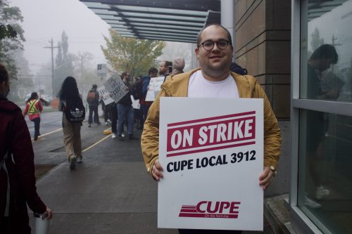 A man smiling at the camera, he is a holding a sign that says On Strike CUPE Local 3912, and standing in front of the strike.