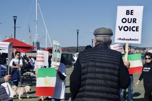 A man holding a sign that says be our voice. Behind him, there are other protestors holding the Iranian flags and posters speaking against Iran's current regime.
