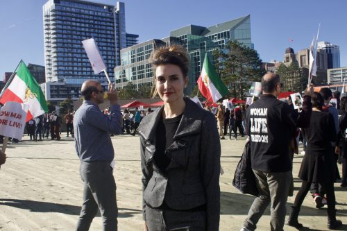 A woman in a suit smiling to the camera, she is seen standing behind an Iranian rally taking place. She is at the Halifax waterfront and people behind her are holding signs calling for the dismantling of the Iranian regime.