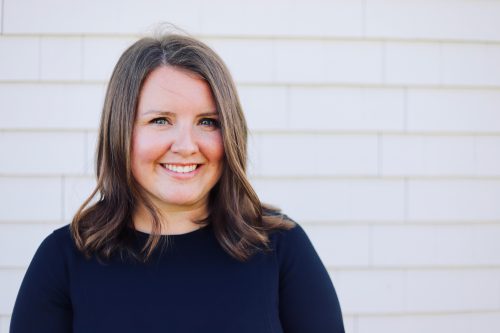 Photo of a woman smiling, standing behind a white background. She is wearing a black shirt.