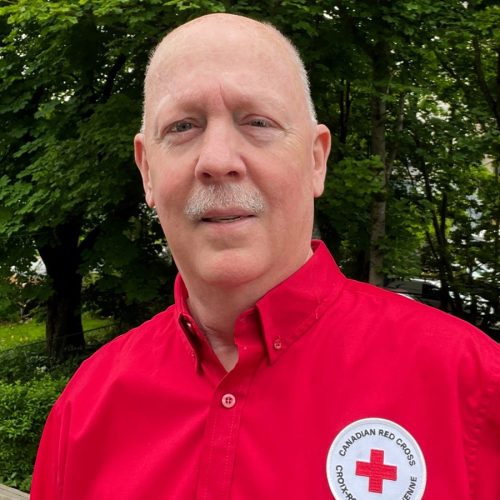 Portrait photo of a man smiling, he is wearing a red shirt that say Canadian Red Cross on it and there are trees in the background.