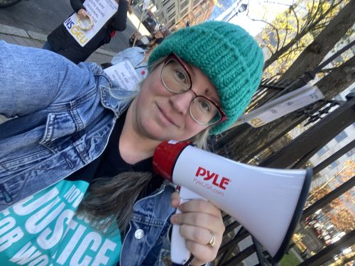 A woman holds a microphone. She is wearing glasses, a jeans jacket and a green beanie and is seen standing on the street in what appears to be a rally.