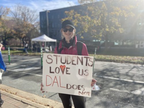 A woman holding a sign that says "students love us Dal does not," she is standing in front of Dalhousie's law building. 