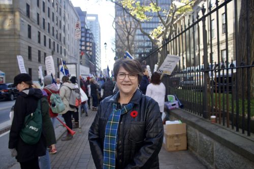 A woman smiling and looking at the camera. She is standing in front of the Nova Scotia Legislature and people are seen rallying in the background. 