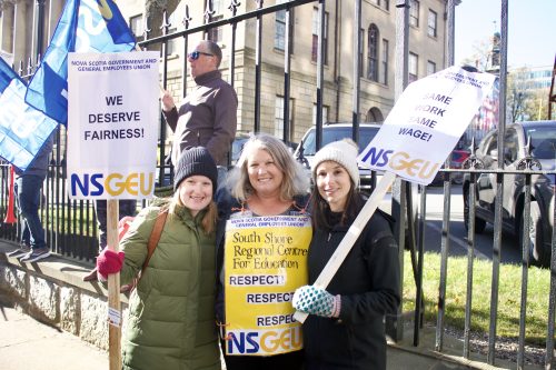 Photo of three women holding signs that say 'We deserve fairness' are seen smiling for the camera. Behind them is the Nova Scotia Legislature building.