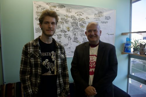 Photo of two men standing and smiling for the photo. Behind them is a couch and drawings on the wall. They are in CKDU, Dalhousie's radio station offices. 