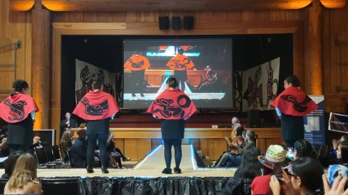 Four women with their backs to the audience showing short red capes that feature traditional indigenous designs.