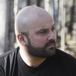 A professional headshot of author SP Joseph Lyons. He looks to the right, he is bald and wearing a brown T-shirt and has a beard.