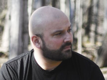 A professional headshot of author SP Joseph Lyons. He looks to the right, he is bald and wearing a brown T-shirt and has a beard.