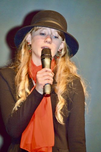 Une jeune fille portant un gilet noir et un chapeau en train de chanter.