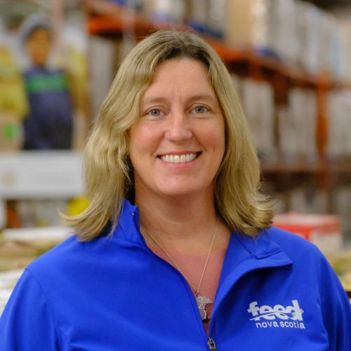 Portrait photo of Karen Theriault, the communications director for Feed Nova Scotia. She is seen smiling and wearing a blue shirt that says Feed Nova Scotia. She is standing in the warehouse, and there are boxes in the background. 