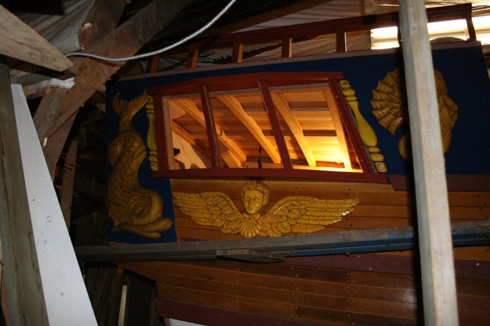 L'extérieur de la cabine du côté triport la poupe du bateaude Jean Belliveau. On voit une fenêtre avec trois cadre. En dessous, une gravure en bois peinturé jaune d'une tête supportée par une coquille avec deux ailles sur ses cotés. À la gauche de la fenêtre, une grande gravure de poisson également peinturée jaune.