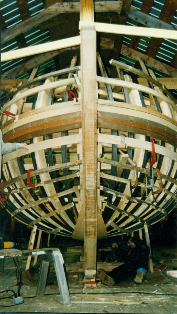 Le cadre avant en bois du navire de Jean Belliveau en construction à l'intérieur d'un atelier. On s'aperçoit d'un homme qui travaille à son bas.
