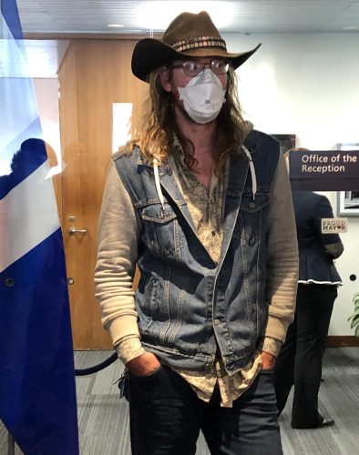 A person with long blonde hair and a cowboy hat. They are standing wearing a denim vest and beige shirt inside an office area.