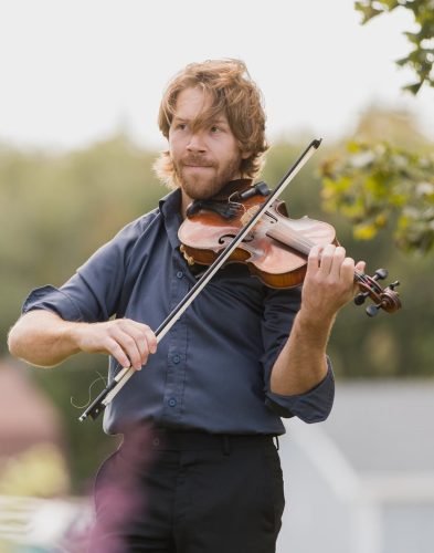 Jacques Blinn jouant du violon par une belle journée d'automne. Le trentenaire porte une chemise et un pantalon formels, a des cheveux châtain clair de longueur moyenne et une courte barbe. 