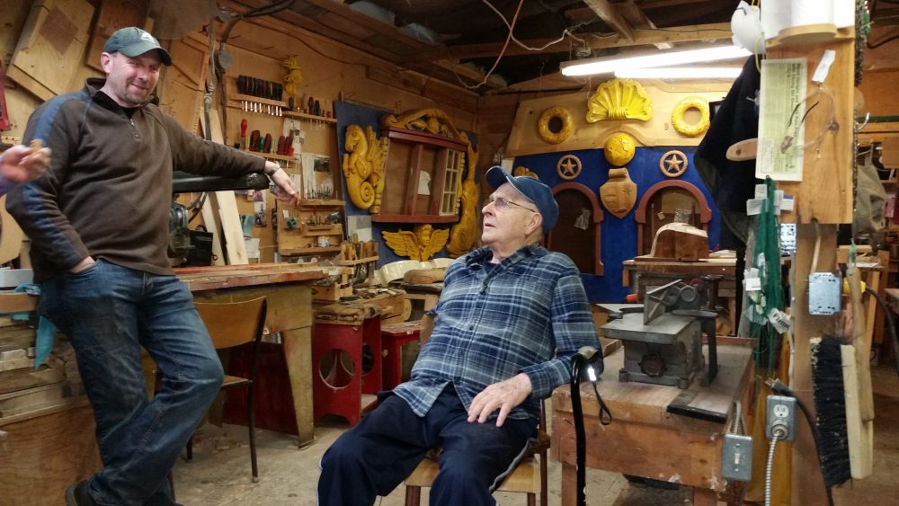 L'ainé Jean Belliveau assis dans son atelier, parlant avec un homme plus jeune. À l'arrière-plan, nous voyons des sculptures en bois pour son bateau.