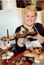 A woman in an apron holds a plate of baked goods and more pastries are in the foreground.