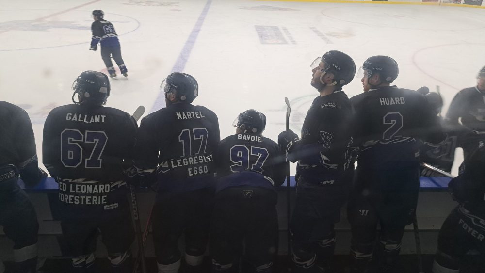 Joueur de hochey en uniforme bleu debout au banc des joueurs.