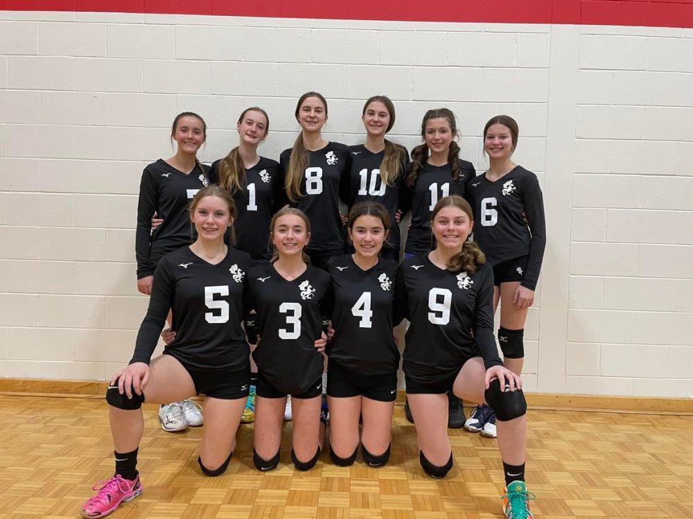 Dix jeunes filles souriantes sont devant un mur de gymnase portant leurs uniformes de volley-ball. Six d'entre-eux sont debouts, et quatre sont à genou devant.
