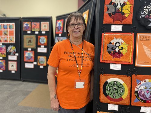 Photo of cultural advisor at the Mi'kmaw Native Friendship Centre Theresa Meuse standing next to the Every Child Matters exhibit. She is wearing an orange shirt with the title Residential Schools on it. 