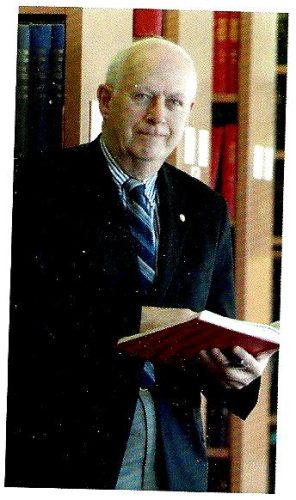 Portrait photo of Dr. Allen Marble, chairman of the Medical History Society of Nova Scotia. He is wearing a suit, smiling and holding a red book in his hand.