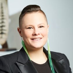 Woman pictured smiling. She's wearing a black blazer and a black shirt and long beaded green earrings,.