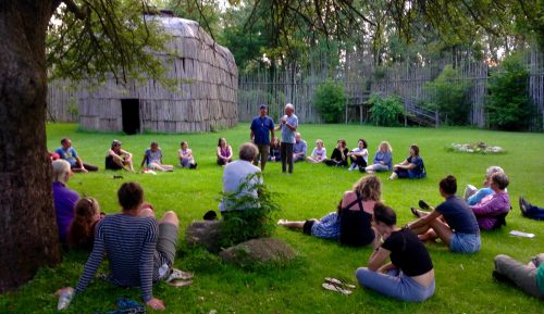 A group of people are pictured sitting in a grassed area with a traditional longhouse in the background. The group of people are surround two men standing in the middle of the group speaking with them. 