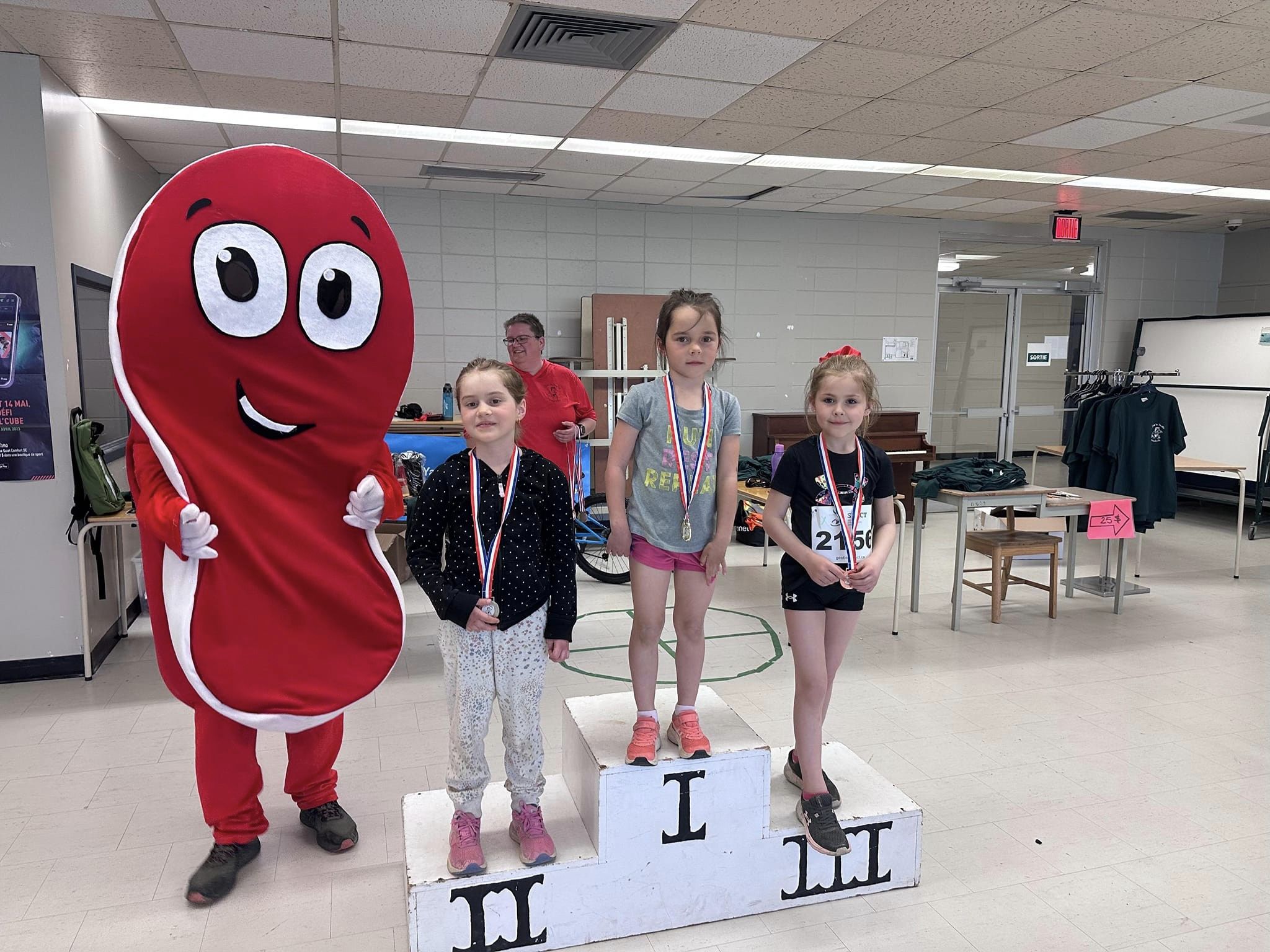 3 filles sur un podium avec une médaille au cou en compagnie d'une mascotte rouge