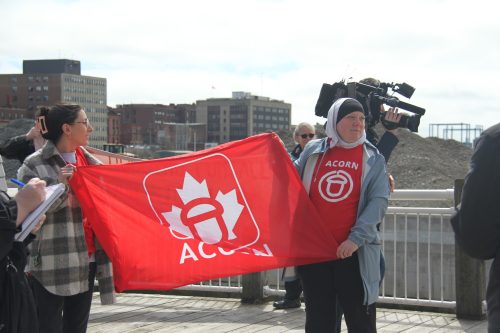 Two people hold a bright red flag for the group ACORN. It includes the word ACORN, written in white, below the image of an acorn superimposed against a maple leaf. A video cameraperson is visible in the background, along with the Saint John cityscape.