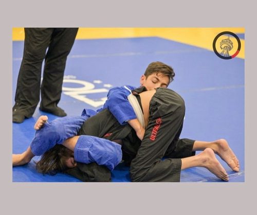 A referee stands at the edge of a blue mat on which DiNola is entangled with a competitor.