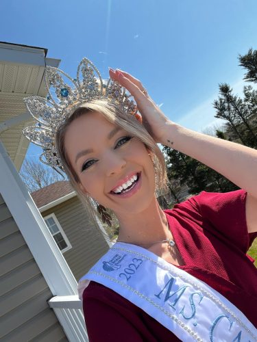 A woman wearing a red shirt, white sash and crown stands outside of a sunny day. She is smiling.