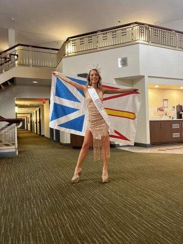 A woman wearing a gold dress holds the flag of Newfoundland in a lobby. 
