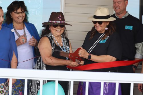 Dianne Meili with people on either side of her, cuts a big red ribbon with a large pair of scissors to mark the grand opening of the cultural center. Weather is clear.