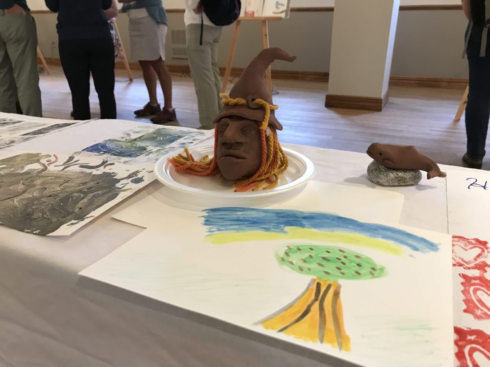 Une sculpture en forme de visage portant un chapeau pointu dans une assisette de carton entourée de plusieurs dessins