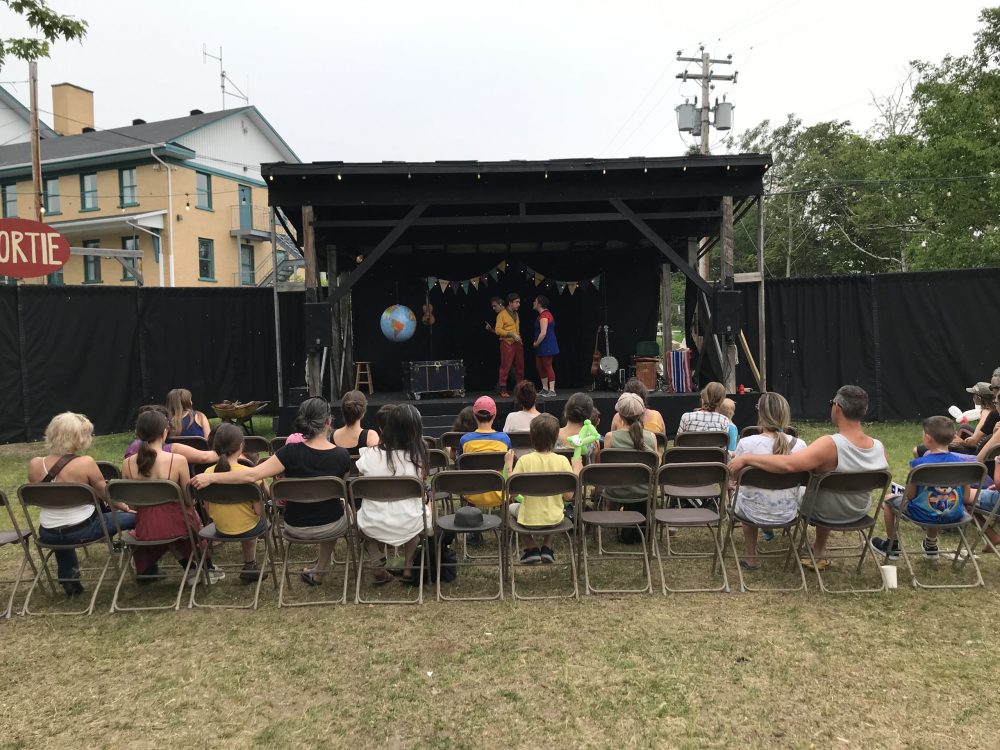 Une foule assise sur des chaises à l'extérieur qui observe les artistes sur la scène