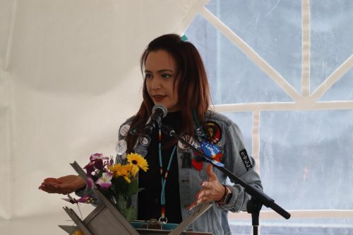 April Eve Wiberg stands at a podium with flowers on them in front of community members sharing stories under a tent along Okîsikow (Angel) Way. Weather outside the tent is cloudy.