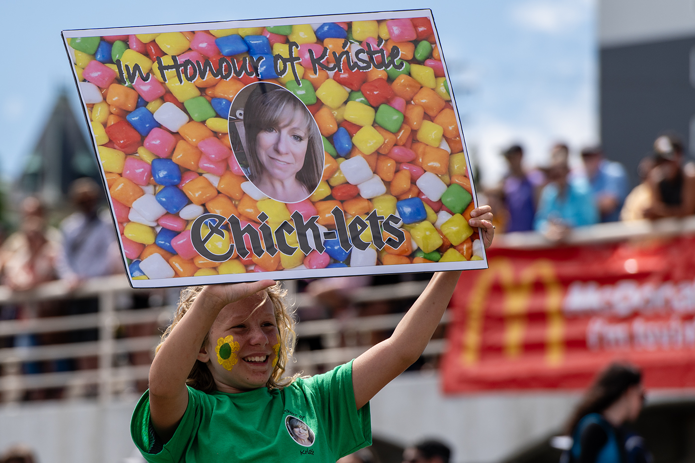 A child with a flower painted on her face smiles and holds up a sign in memory of her mother to a crowd watching the Silly Boat races