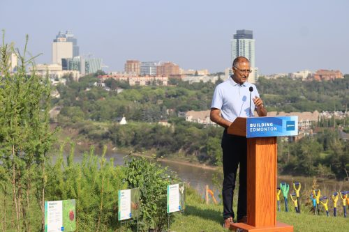 Mayor Amarjeet Sohi doing a press conference on a lookout that has a huge backdrop of the North Saskatchewan River, as well as other high-rises on the other side of the valley. Weather is clear.