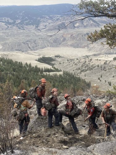 A crew of wildfire firefighters stands overlooking a dry valley, initial attack crew leader Ashley Holyk is looking back and smiling at the camera.