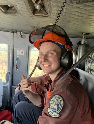 A woman wearing a BC Wildfire Service shirt and wearing an orange hard hat smiles inside a helicopter as it flys over forests. 