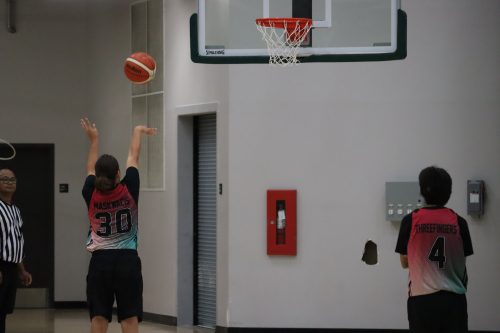 A basketball player shoots a free throw, with a teammate and a referee standing on either side waiting for play to resume. Photo was taken inside. 