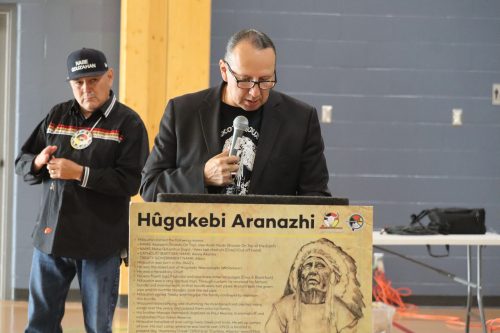 Chief Tony Alexis stands at a podium to share stories in front of his nation, with an organizer standing behind him. The photo was taken in a gymnasium.