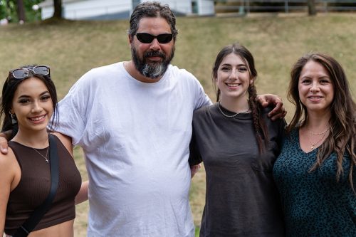 A man in a white t-shirt and wearing sunglasses stands with three women smiling with a hill of brown grass behind them.