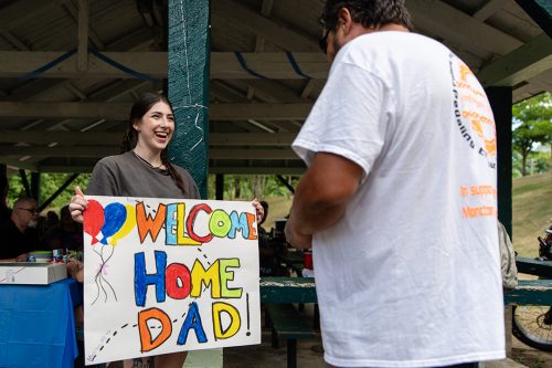 A woman smiles and holds a sign reading Welcome Home Dad as a man in a white shirt reads it. In the background is a group of people eating under a picnic shelter.