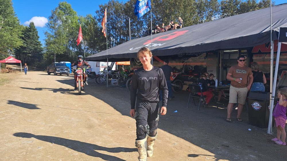 Un garçon de 15 ans debout en habit de motocross noir, lors d'une compétition de motocross pendant une journée ensoleillé