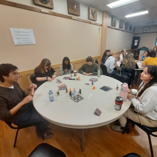 Members of the UNBC Community are gathered around a round table. The 5 people are conversing and folding together origami ravens. Another round table with students seated at it can be seen in the background.
