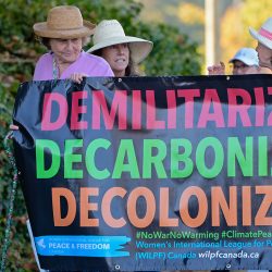 Two protesters hold a banner reading demilitarize, decarbonize, decolonize.