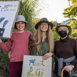 Three generations of a family stand in front of a green background holding signs that read "The oceans are rising and so are we" and "Planet before profit$"