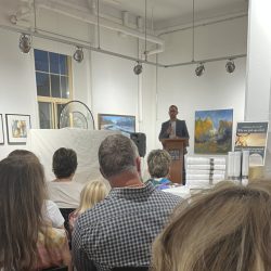 Joseph Gibbons stands before a seated crowd in an art gallery presenting his new book.